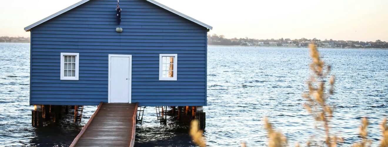 A tranquil scene of a blue boathouse on Swan River, with a wooden walkway and soft lighting.