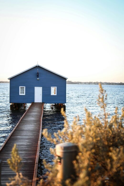 A tranquil scene of a blue boathouse on Swan River, with a wooden walkway and soft lighting.