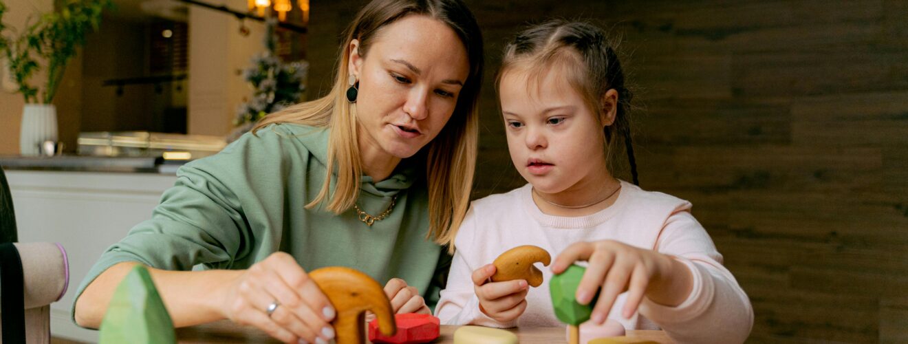 A mother and daughter engaging in play with wooden toys at a table indoors.