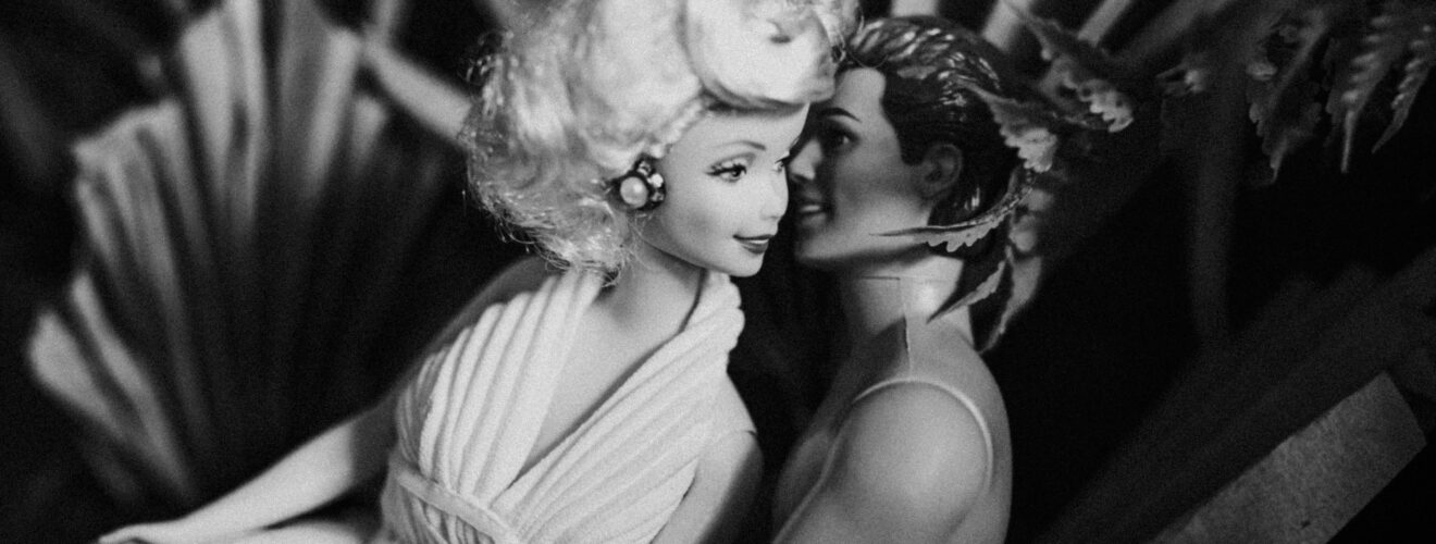 Vintage-style black and white photo of Barbie and Ken dolls in a romantic embrace amidst foliage.