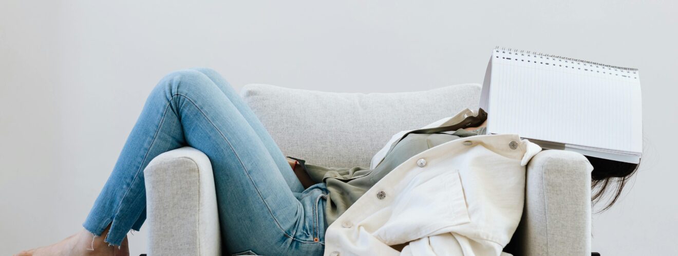 Tired woman in casual attire falls asleep on sofa with a notebook on face and pen in hand.